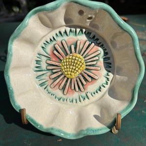 May include: A ceramic dish with a floral design. The dish has a cream-colored base with a turquoise rim and a raised flower in the center. The flower has a yellow center, pink petals, and green accents.