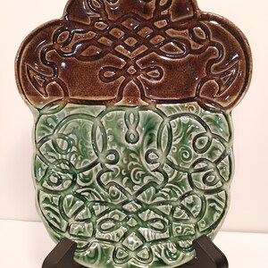 May include: A decorative ceramic plate with a brown top section and a green bottom section. The plate features an intricate Celtic knot design. The plate is displayed on a black stand.