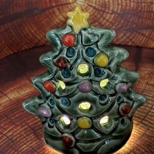May include: A ceramic Christmas tree figurine in a glossy sage green glaze, featuring a yellow star topper and multi-colored raised circular ornaments in red, blue, and yellow. The structure has cut-out openings that emit a warm internal light.