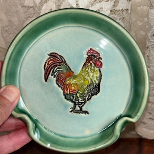 May include: A round ceramic spoon rest featuring a light blue-green glazed surface with a raised, hand-painted rooster in the center. The rooster displays vibrant red, yellow, and black plumage. The spoon rest has a dark green rim and a notched edge.