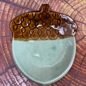 May include: A ceramic dish shaped like an acorn with a glossy finish. The top cap section is dark brown with a textured, looped pattern, while the bottom nut section is a smooth, pale mint green color with subtle speckling throughout.