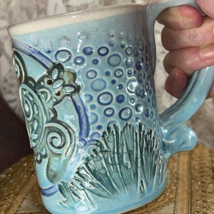 May include: A handmade ceramic mug with a light blue glaze. The mug features textured patterns of bubbles and stylized aquatic plants. The handle and rim are a lighter shade of blue and beige. The mug is held by a person.