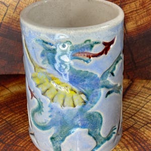 May include: A ceramic mug with a blue dragon design. The dragon is depicted with blue, yellow, and red accents. The mug has a light blue background and a handle. The mug is cylindrical and appears to be handmade.