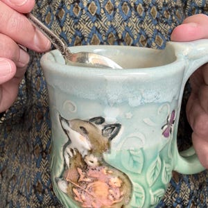 May include: A light green ceramic mug featuring a raised, sculpted fox figure with a pink floral accent. The mug has a smooth, glossy finish with a lighter, pale green rim and a textured leaf pattern around the base. A silver spoon rests inside.