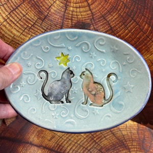 May include: An oval ceramic dish in light blue with a glossy finish. The center features two embossed cats, one grey and one peach-colored, facing each other. A single yellow star is positioned above them, surrounded by embossed swirls and stars.