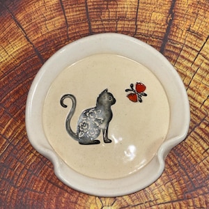 May include: A ceramic spoon rest with a cream-colored interior and a white rim. The rest features a black cat with floral detailing and a red and black butterfly. The spoon rest has a curved shape with a small handle on each side.
