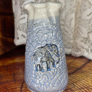 May include: A ceramic vase with a light blue and white color scheme. The vase features a textured surface with an embossed elephant design. The top of the vase has a ruffled edge.