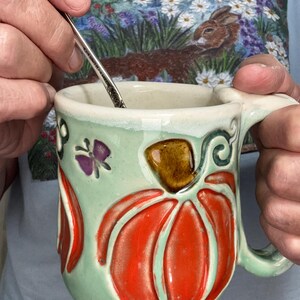 May include: A ceramic mug with a light green glaze featuring a raised, orange pumpkin design with a brown stem and a small purple butterfly accent. A silver-toned metal spoon is inside the mug, which has a curved handle and a textured surface.