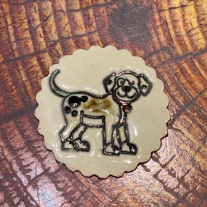 May include: A round, scalloped-edge ceramic pin featuring a glossy cream-colored surface with a black-outlined illustration of a spotted dog. The dog has a small red collar, and the design is finished with a smooth, reflective glaze.