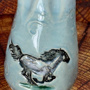 May include: A light blue ceramic vessel with a glossy finish, featuring a raised, dark-toned relief of a galloping horse. The surface is decorated with subtle, swirling embossed patterns that catch the light, creating a textured appearance.