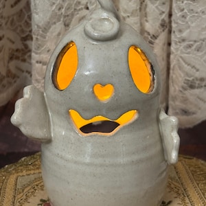 May include: A speckled gray ceramic ghost-shaped candle holder with a curly top and small arms. The ghost has cut-out eyes, a heart-shaped mouth, and a glowing orange light inside. The candle holder is a Halloween decoration.