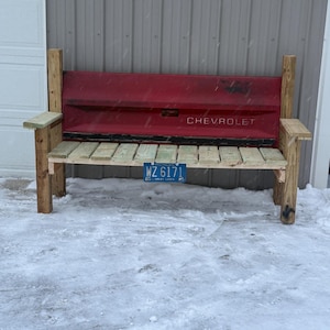 May include: A rustic bench made from a red Chevrolet truck tailgate, with the word "CHEVROLET" visible. The seat is constructed from wooden planks, and the bench has wooden armrests and legs. A license plate is attached to the front.
