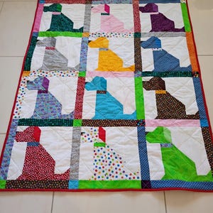 May include: A rectangular patchwork quilt featuring a grid of twelve dog silhouettes in various colors and patterns, including green, pink, purple, gray, yellow, blue, brown, red, and polka-dot fabrics, all set against white backgrounds with a red border.