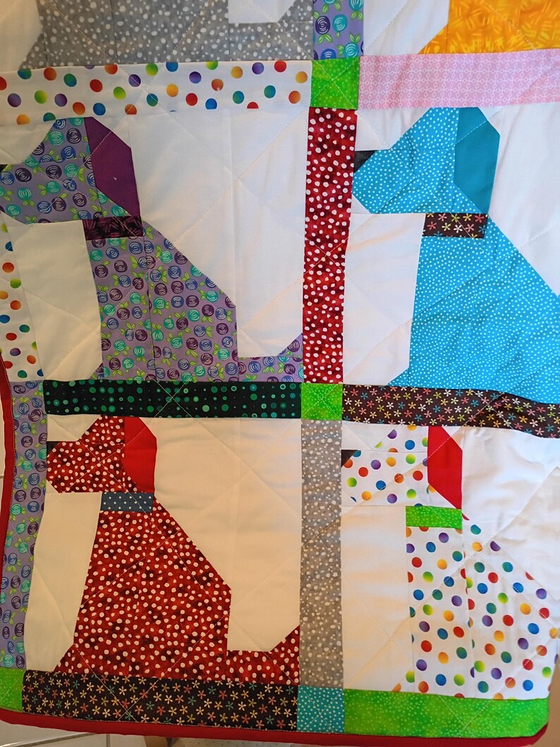 May include: A patchwork quilt featuring four stylized dog silhouettes crafted from various colorful fabrics. The dogs are rendered in purple, blue, red, and white with polka dot, floral, and geometric patterns, separated by white quilted sections.