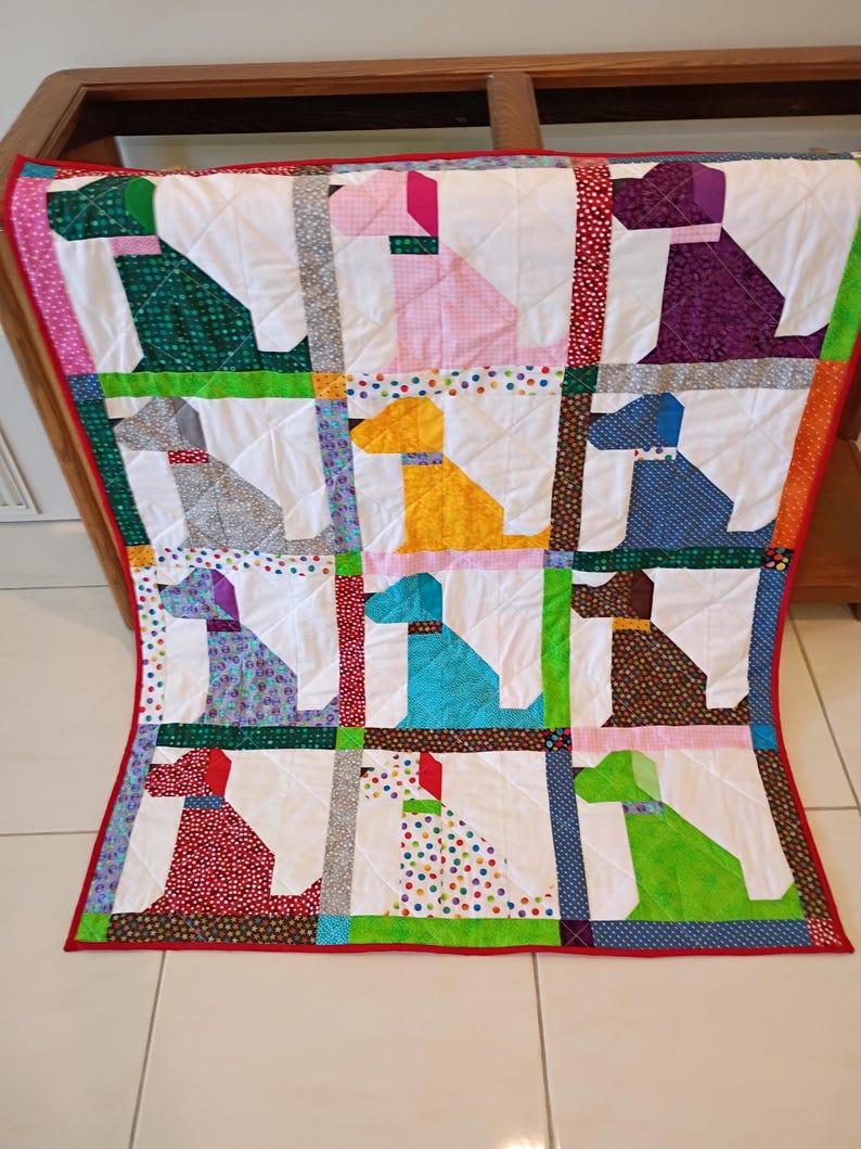 May include: A patchwork quilt featuring a grid of twelve stylized dog silhouettes in various colors and patterns, including green, pink, purple, gray, yellow, blue, brown, red, and polka dots, separated by white fabric and a red border.