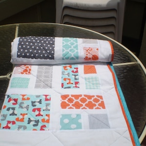 May include: A handmade baby quilt with a fox pattern in orange, teal, and gray. The quilt features various geometric patterns and is bordered with orange and teal trim. The rolled-up quilt on top has a gray triangle pattern.