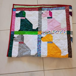 May include: A square patchwork quilt featuring four stylized dog silhouettes in green, pink, gray, and yellow. The quilt is constructed with white background blocks and separated by colorful patterned fabric strips, finished with a red border.