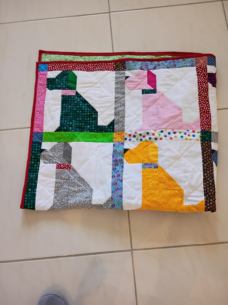 May include: A square patchwork quilt featuring four stylized dog silhouettes in green, pink, gray, and yellow. The quilt is constructed with white background blocks and separated by colorful patterned fabric strips, finished with a red border.
