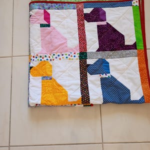 May include: A square quilted blanket featuring four dog silhouettes in pink, purple, yellow, and blue patterned fabrics. The quilt has white background blocks, colorful polka dot and floral sashing, and a bright red border with a green backing.