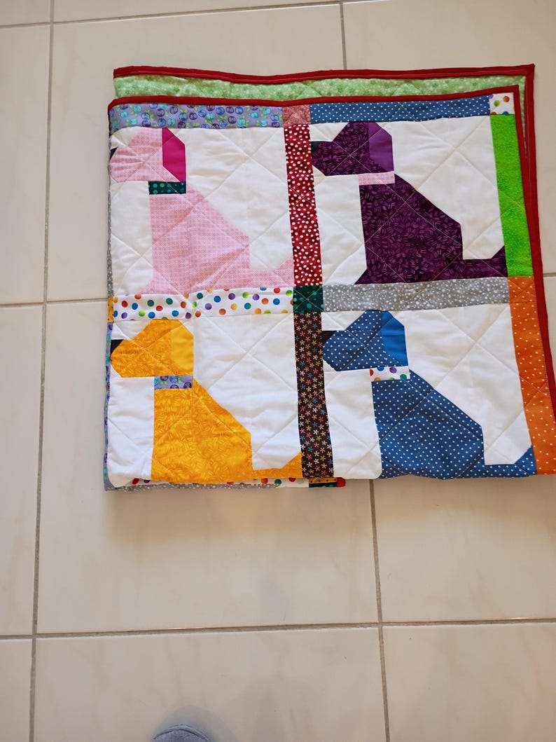 May include: A square quilted blanket featuring four dog silhouettes in pink, purple, yellow, and blue patterned fabrics. The quilt has white background blocks, colorful polka dot and floral sashing, and a bright red border with a green backing.