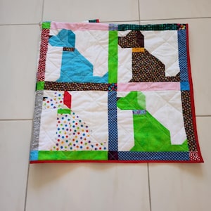 May include: A square patchwork quilt featuring four distinct dog silhouettes in blue, brown, white with colorful polka dots, and green. The quilt is constructed with white fabric blocks, separated by patterned fabric sashing, and finished with a red border.