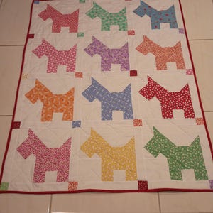 May include: A rectangular quilt featuring a white background with a grid of twelve fabric dog silhouettes in various colors and patterns, including pink, green, blue, purple, orange, and yellow, finished with a solid red fabric border.