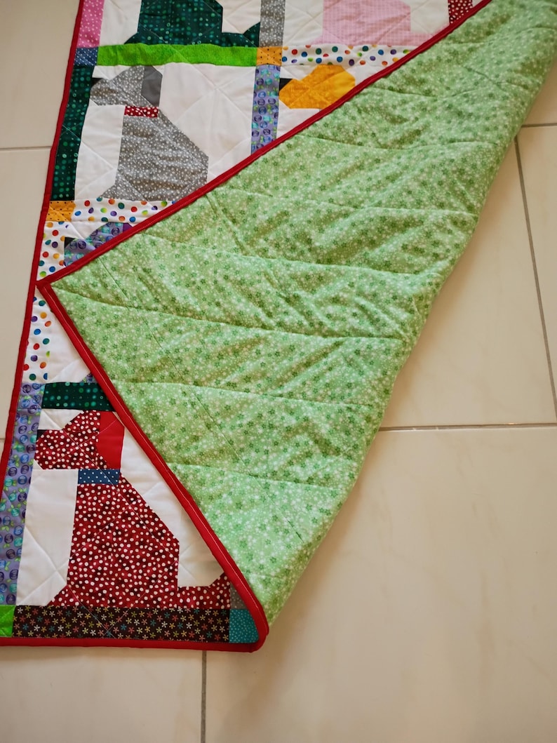 May include: A rectangular patchwork quilt featuring stylized dog silhouettes in various patterned fabrics including red, grey, and yellow. The quilt has a bright red border and a light green backing fabric with a small white floral print pattern.