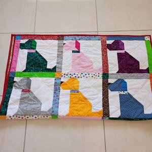 May include: A rectangular quilted blanket featuring a grid of six dog silhouettes in green, pink, purple, gray, yellow, and blue patterned fabrics. The quilt is bordered by a red fabric edge and includes colorful polka dot and floral fabric sashing.
