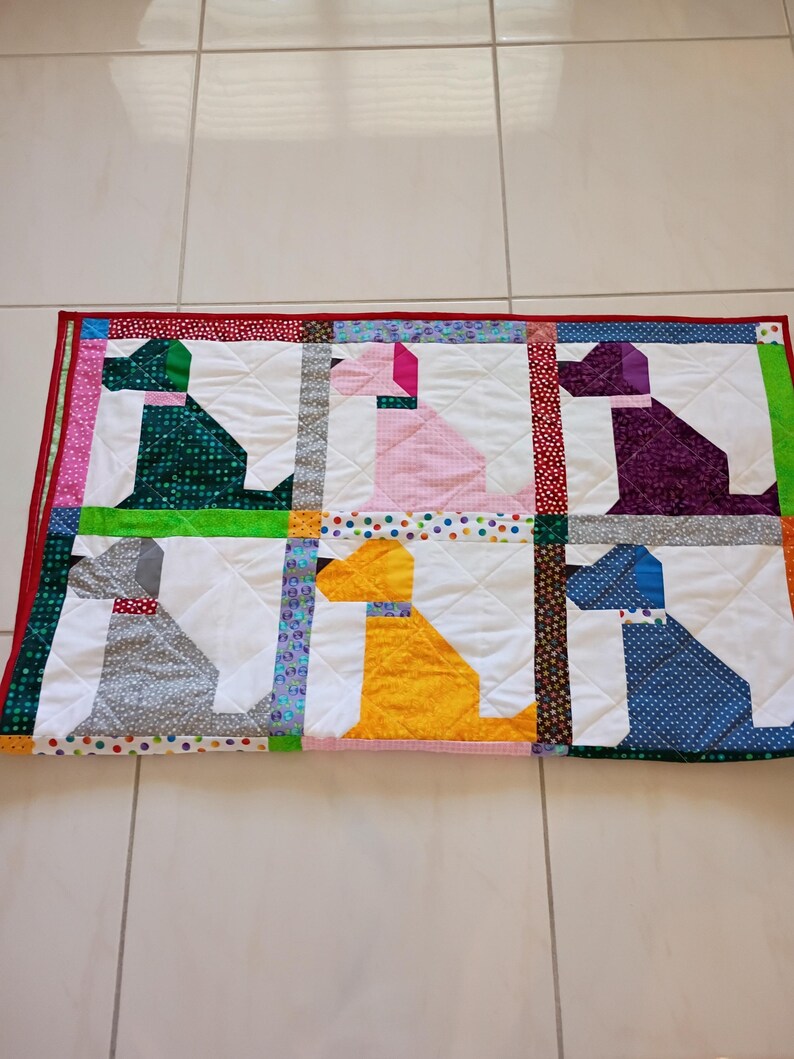 May include: A rectangular quilted blanket featuring a grid of six dog silhouettes in green, pink, purple, gray, yellow, and blue patterned fabrics. The quilt is bordered by a red fabric edge and includes colorful polka dot and floral fabric sashing.