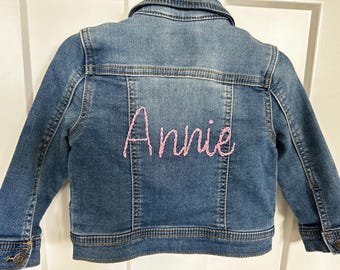 Könnte beinhalten: Hellblaue Jeansjacke mit dem Namen "Annie" in rosa Faden auf dem Rücken gestickt. Die Jacke hat ein klassisches Design mit Kragen, Knopfverschlüssen und Taschen.