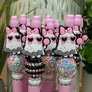 May include: A collection of pink pens with decorative toppers. Each pen features a ghost-shaped charm wearing heart-shaped sunglasses and a bow. The pen barrels are pink, and the toppers are embellished with rhinestones and pink accents.