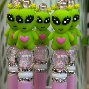 May include: A collection of pink pens topped with green alien figures. Each alien has a pink heart, black eyes, and yellow star accents. The pen bases have a clear glitter globe and decorative silver accents.