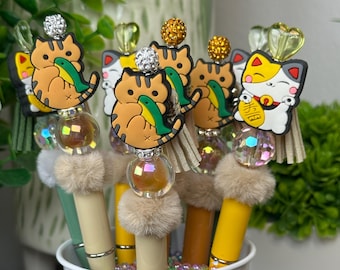 May include: Five colorful pens with cat-themed toppers. The toppers are made of plastic and feature a cat holding a fish. The pens have a yellow and green body with a fluffy white pom-pom at the bottom. The pens are in a white bucket.