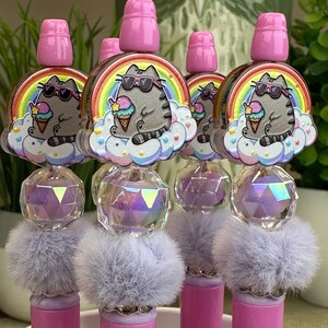 May include: A collection of pink and gray pens. Each pen features a cartoon cat wearing sunglasses and holding ice cream, set against a rainbow and cloud background. The pens have crystal and fluffy accents.
