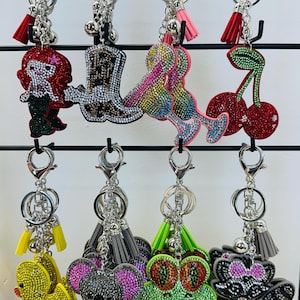 May include: A collection of rhinestone-studded keychains featuring various designs. The keychains include a mermaid, cowboy boot, cocktail glasses, cherries, a duck, a koala, a frog, and a raccoon. Each keychain has a silver-tone ring, a tassel, and a small bell.