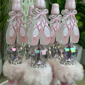May include: A collection of decorative bottle stoppers. Each stopper features a pink ballet slipper design with a bow, a clear iridescent orb, and a fluffy pink base. The stoppers are topped with a light pink cap.