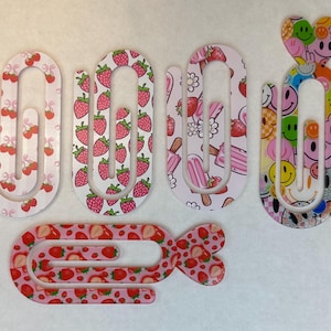 May include: A collection of decorative paper clips with various patterns. Designs include strawberries, cherries, ice cream, and smiley faces. The clips are in shades of pink, red, white, and other colors. The clips are made of a clear material.