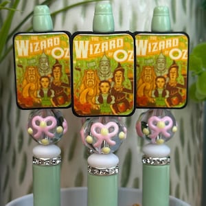 May include: Three decorative bottle stoppers featuring a "Wizard of Oz" design. Each stopper has a light green base, a clear bead with a pink bow, and a rectangular top with the movie's characters.