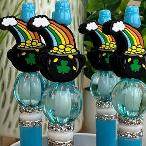 May include: A collection of pens with a St. Patrick's Day theme. Each pen features a black pot of gold charm with a rainbow and shamrock design. The pens have a blue barrel, a clear blue orb, and a blue top.