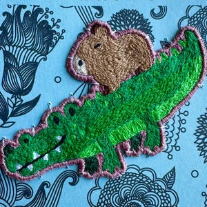 May include: A light blue card with the words "THANK YOU" in black. The card features an embroidered green alligator with a brown bear on its back. The card has a floral pattern and the Etsy shop name.