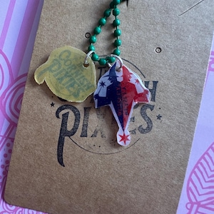 Puede incluir: Un collar de cuentas verdes con dos colgantes. Un colgante es una forma amarilla translúcida con las palabras "Cornish Pixies". El otro colgante es un diseño rojo, blanco y azul con una estrella. Los colgantes están unidos a una etiqueta marrón.