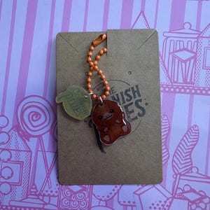 Puede incluir: Un llavero marrón con una cadena naranja y un dije de ornitorrinco de dibujos animados. El dije está sujeto a un respaldo de cartón con el texto "The Cornish Pixies". El fondo es un patrón de rayas rosas y blancas.