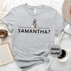K&ouml;nnte beinhalten: Graues T-Shirt mit einem Cartoon-Bild von Olaf aus Frozen, der &uuml;ber eine Linie schaut, unter der der Text "SAMANTHA?" gedruckt ist.