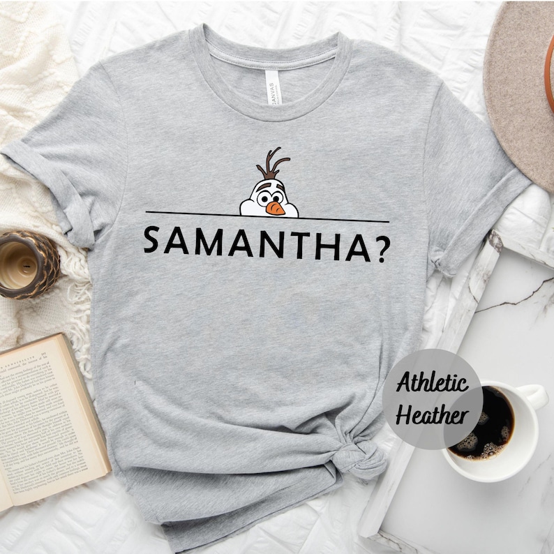 K&ouml;nnte beinhalten: Graues T-Shirt mit einem Cartoon-Bild von Olaf aus Frozen, der &uuml;ber eine Linie schaut, unter der der Text "SAMANTHA?" gedruckt ist.