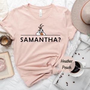 K&ouml;nnte beinhalten: Ein pinkfarbenes T-Shirt mit einem Cartoon-Bild von Olaf aus Frozen, der &uuml;ber eine schwarze Linie mit dem Text "SAMANTHA?" schaut. Das Shirt ist aus weichem, heidefarbenem Pfirsichstoff gefertigt.