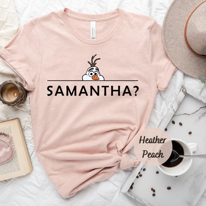 K&ouml;nnte beinhalten: Ein pinkfarbenes T-Shirt mit einem Cartoon-Bild von Olaf aus Frozen, der &uuml;ber eine schwarze Linie mit dem Text "SAMANTHA?" schaut. Das Shirt ist aus weichem, heidefarbenem Pfirsichstoff gefertigt.
