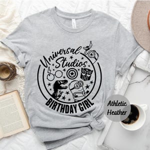 May include: Grey t-shirt with a black graphic of a circle with the text "Universal Studios Birthday Girl" and images of a minion, a dinosaur, a pair of glasses, a star, a shield, and a robot. The text "Athletic Heather" is printed in black on the bottom right corner of the shirt.