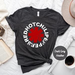 May include: Dark grey heather t-shirt featuring a distressed white graphic of the words "Red Hot Chili Peppers" encircling a red asterisk. The shirt is made of soft material and is suitable for casual wear. The shirt is knotted at the bottom.