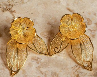 May include: A pair of gold-colored filigree flower earrings with leaf accents. The earrings are displayed on a marble-like surface, with dried foliage in the background. The earrings have a delicate, openwork design.