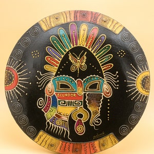May include: A round, black wooden wall hanging with a colorful, intricate design. The design features a stylized face with a butterfly above it, and the word "Ecuador" written in the center. The design is surrounded by a border of gold and red.